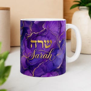 Puede incluir: Taza de cerámica blanca con un vibrante diseño abstracto en morado y dorado. La taza presenta el nombre "Sarah" en letras doradas, con texto hebreo encima. La taza tiene un asa blanca y es adecuada para bebidas frías o calientes.