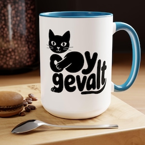 May include: White ceramic mug with a blue interior and handle. A black cat graphic is featured on the front, along with the text "oy gevalt" in a playful font. The mug is on a wooden surface.