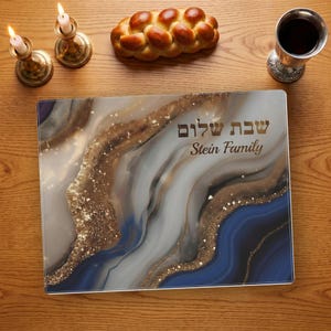 May include: A rectangular placemat featuring a marbled pattern in shades of blue, white, gray, and gold glitter. The text "Shabbat Shalom" in Hebrew and "Stein Family" in English are printed in gold on the right side of the placemat surface.