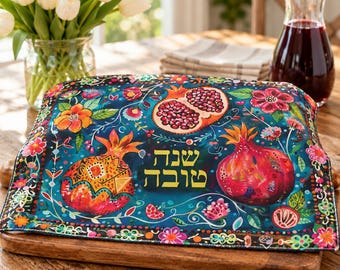 May include: A rectangular fabric challah cover featuring a dark blue background with colorful illustrations of pomegranates, flowers, and leaves. The Hebrew text "Shana Tova" is printed in yellow in the center of the fabric.