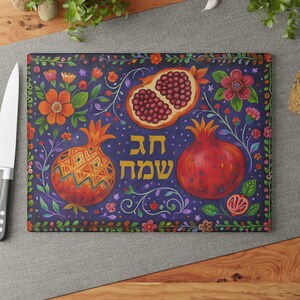 May include: A rectangular glass cutting board featuring a dark purple background with colorful illustrations of pomegranates and flowers. The Hebrew text "חג שמח" is printed in yellow in the center. A white kitchen knife is positioned on the left side.