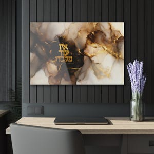 Puede incluir: Impresión artística abstracta con texto hebreo dorado sobre un fondo de colores marrones, dorados y blancos arremolinados. La obra de arte se exhibe en un escritorio con un portátil y un jarrón de lavanda.