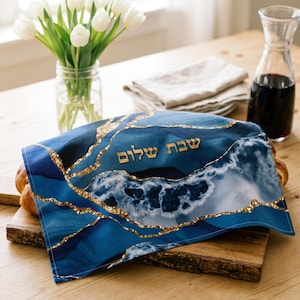 May include: A rectangular fabric challah cover featuring a deep blue and white marbled pattern with gold-colored veins. The Hebrew text "Shabbat Shalom" is embroidered in gold lettering across the center of the fabric.