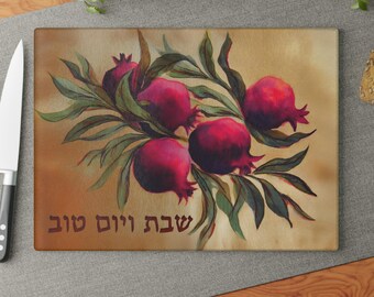 May include: A rectangular glass cutting board featuring a painted illustration of deep red pomegranates with green leaves on a tan background. The Hebrew text "שבת ויום טוב" is printed in dark brown lettering at the bottom of the board.