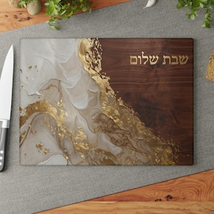 May include: A rectangular cutting board featuring a dark brown wood grain section on the right and a white, gray, and gold marbled resin design on the left. The Hebrew text "שבת שלום" is printed in gold on the wood portion of the board.