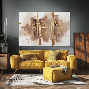 May include: Abstract art canvas with gold Hebrew script over a background of brown and beige brushstrokes. The artwork hangs above a yellow sofa with matching ottoman and throw pillows. The room has a modern, minimalist design.
