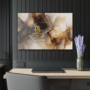Puede incluir: Una impresión artística abstracta con texto hebreo dorado sobre un fondo de colores marrones, beige y blancos arremolinados. La obra de arte se exhibe en una pared sobre un escritorio con un portátil y un jarrón de lavanda.