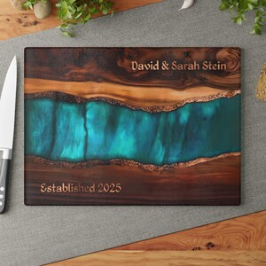 May include: A rectangular wooden cutting board featuring a central river-like inlay of translucent teal resin with copper-colored edges. The wood has dark brown grain patterns. Engraved text reads David & Sarah Stein and Established 2025.