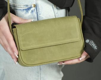 May include: A rectangular olive green crossbody bag with a smooth matte finish and a fold-over flap closure. The bag features a matching thin shoulder strap and visible surface texture with subtle natural markings across the front panel.