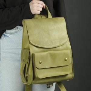 May include: A sage green leather backpack featuring a large top flap, a prominent front pocket with two brass-colored snap buttons, and a side pocket with a matching snap closure. The bag includes a top handle and adjustable shoulder straps.
