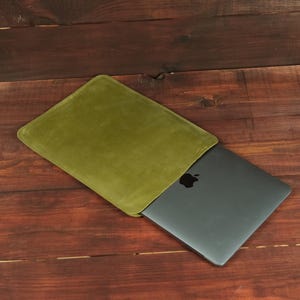 Peut inclure: Une housse pour ordinateur portable rectangulaire de couleur vert olive avec une finition lisse, contenant partiellement un ordinateur portable gris foncé avec un logo pomme noir visible sur le couvercle. La housse possède une ouverture latérale.