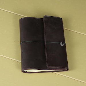 Pode incluir: Uma capa de di&aacute;rio em couro roxo escuro com um acabamento liso e mate. A capa &eacute; fixada por uma fina banda el&aacute;stica preta que envolve horizontalmente o centro, apresentando um pequeno detalhe de conta met&aacute;lica prateada na parte frontal.