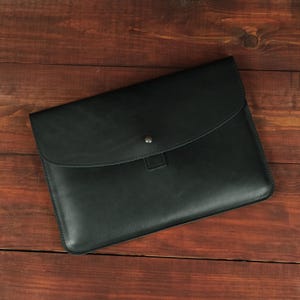May include: A black leather document sleeve featuring a smooth, matte finish and a curved flap closure. The envelope-style case is secured with a single centered metal snap button, providing a clean, structured appearance for storing papers.
