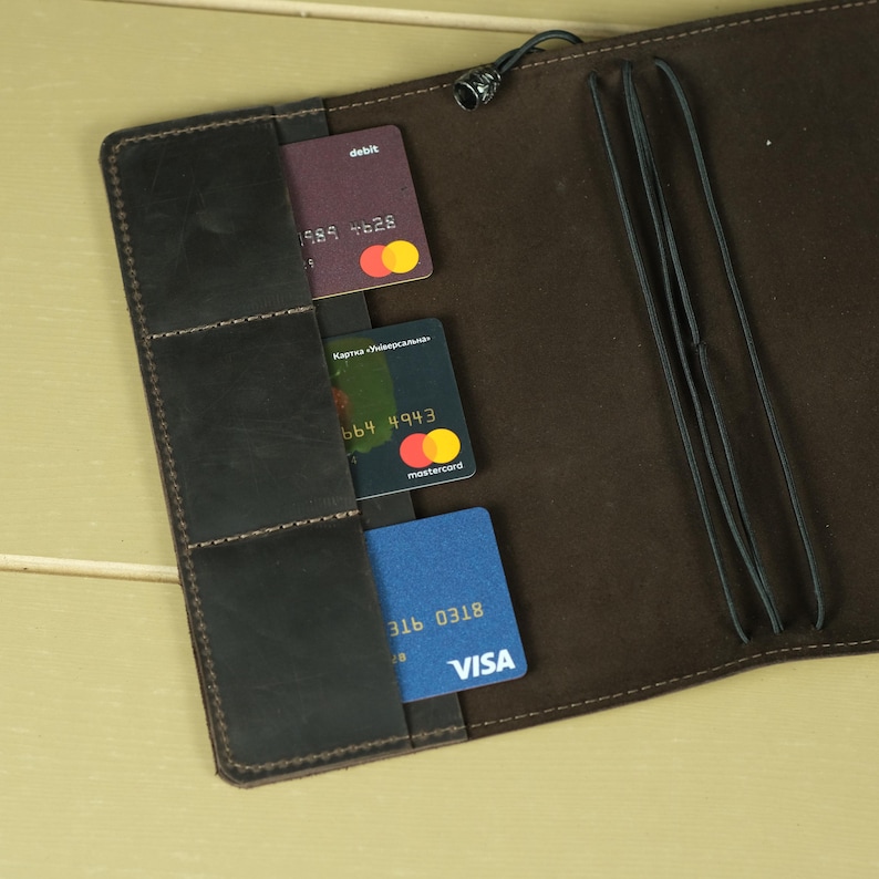 Pode incluir: Um organizador de couro castanho escuro com tr&ecirc;s ranhuras verticais para cart&otilde;es contendo um cart&atilde;o de d&eacute;bito roxo, um Mastercard verde escuro e um cart&atilde;o Visa azul. O interior inclui el&aacute;sticos pretos para prender itens.