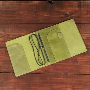 May include: A light green leather notebook cover featuring a matte finish, a vertical zippered pocket, a small pen loop, and two black elastic cords for securing inserts. The interior layout includes two side flaps for holding paper covers.