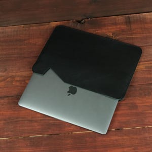 May include: A dark gray laptop with a visible Apple logo partially inserted into a black sleeve. The sleeve features a diagonal cutout design on the front, revealing the metallic surface of the laptop casing beneath the black material.