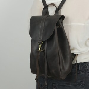 Pu&ograve; includere: Uno zaino in pelle marrone scuro con chiusura a patta ripiegabile fissata da un moschettone in metallo color ottone. La borsa include spallacci sottili e una maniglia superiore, con cuciture visibili lungo i bordi della patta e del corpo.