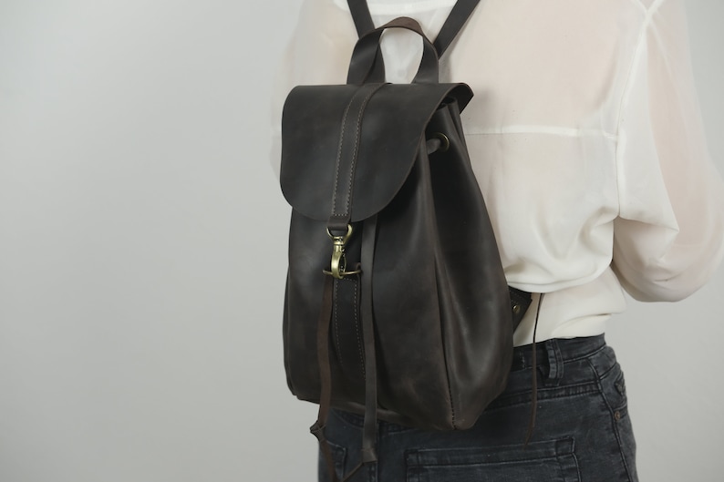 Pu&ograve; includere: Uno zaino in pelle marrone scuro con chiusura a patta ripiegabile fissata da un moschettone in metallo color ottone. La borsa include spallacci sottili e una maniglia superiore, con cuciture visibili lungo i bordi della patta e del corpo.