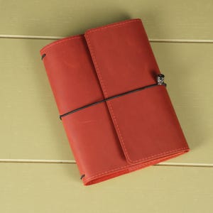 Pode incluir: Uma capa de di&aacute;rio em couro vermelho com acabamento fosco liso, apresentando um fecho de cord&atilde;o el&aacute;stico preto vertical que envolve o objeto e &eacute; fixado por uma pequena conta met&aacute;lica. A capa retangular tem cantos arredondados e costuras.