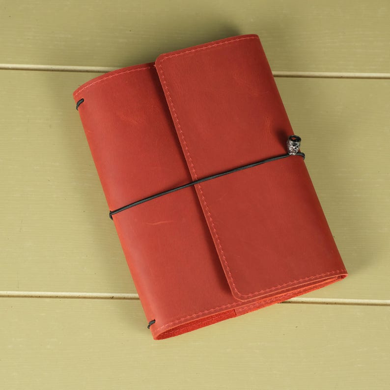 Pode incluir: Uma capa de di&aacute;rio em couro vermelho com acabamento fosco liso, apresentando um fecho de cord&atilde;o el&aacute;stico preto vertical que envolve o objeto e &eacute; fixado por uma pequena conta met&aacute;lica. A capa retangular tem cantos arredondados e costuras.