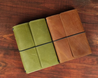 May include: Two leather journals, one in olive green and one in tan brown, each featuring a smooth matte finish and a vertical black elastic band wrapped around the center to secure the covers closed. The journals are rectangular in shape.