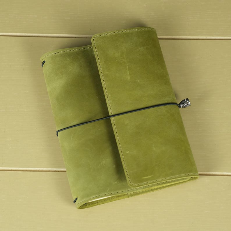 Pode incluir: Uma capa de di&aacute;rio em couro verde-lim&atilde;o com acabamento fosco e costuras vis&iacute;veis ao longo das bordas. A capa &eacute; presa por um cord&atilde;o el&aacute;stico preto fino que envolve horizontalmente o centro, apresentando um pequeno pingente met&aacute;lico no fecho.
