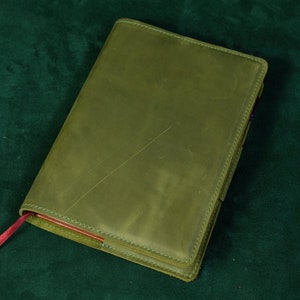 May include: A rectangular notebook with a matte olive green leather cover featuring visible surface scratches and stitched edges. A thin, flat red ribbon bookmark extends from the bottom left corner, contrasting with the muted green exterior.