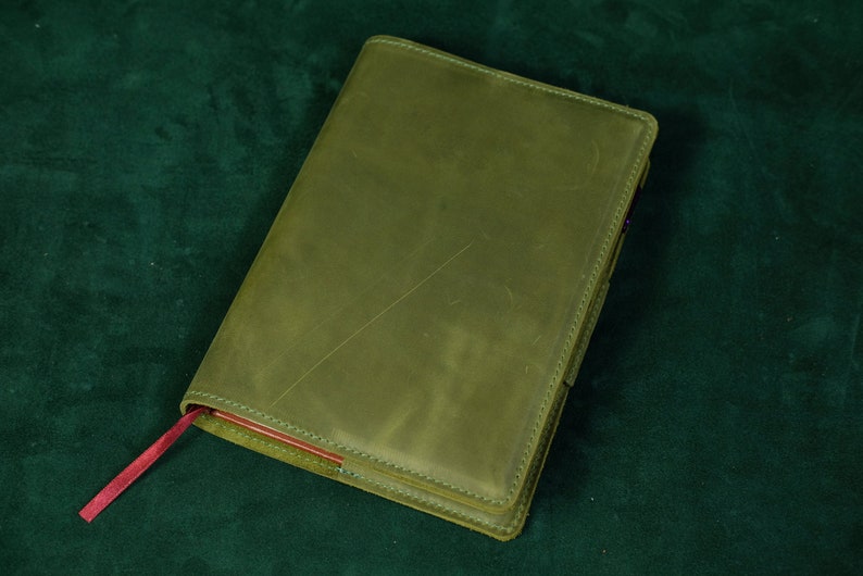 May include: A rectangular notebook with a matte olive green leather cover featuring visible surface scratches and stitched edges. A thin, flat red ribbon bookmark extends from the bottom left corner, contrasting with the muted green exterior.