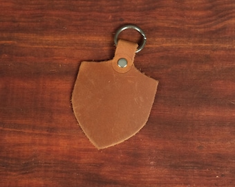 May include: A shield-shaped brown leather keychain featuring a smooth, matte surface texture. The leather piece is attached to a circular silver-toned metal split ring by a small leather tab secured with a single silver-colored metal rivet at the top.