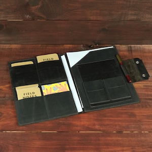 May include: A black leather folio organizer featuring multiple interior pockets, a vertical slot for a notepad, and a pen loop. The left side holds two Field Notes notebooks and a patterned card, while the right side contains a white paper sheet.