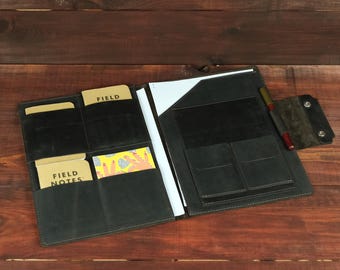 May include: A black leather folio organizer featuring multiple interior pockets, a vertical slot for a notepad, and a pen loop. The left side holds two Field Notes notebooks and a patterned card, while the right side contains a white paper sheet.