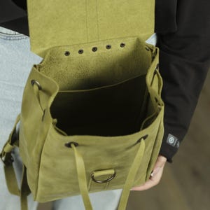 Pu&ograve; includere: Uno zaino in pelle verde oliva con patta ripiegabile, occhielli in metallo scuro e chiusura a coulisse. La borsa presenta un anello a D centrale sul pannello frontale, spallacci regolabili e uno scomparto principale aperto con cuciture.