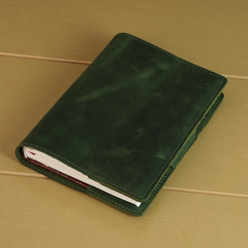 May include: A rectangular notebook featuring a dark forest green cover with a smooth, matte texture and visible tonal variations. The cover is stitched along the edges with matching green thread, enclosing a stack of white paper pages inside.