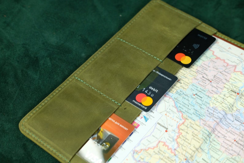 May include: An olive green leather passport or document holder featuring multiple interior card slots. The slots contain two black Mastercard debit cards and one orange Visa card. A detailed map of Ukraine is visible tucked into the holder.