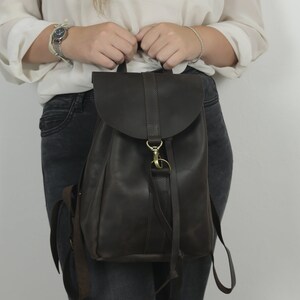 Pu&ograve; includere: Uno zaino in pelle marrone scuro con chiusura a patta ripiegabile fissata da un gancio in metallo color ottone e una chiusura ad anello. La borsa include spallacci regolabili e una maniglia superiore, con una finitura liscia e opaca esterna.