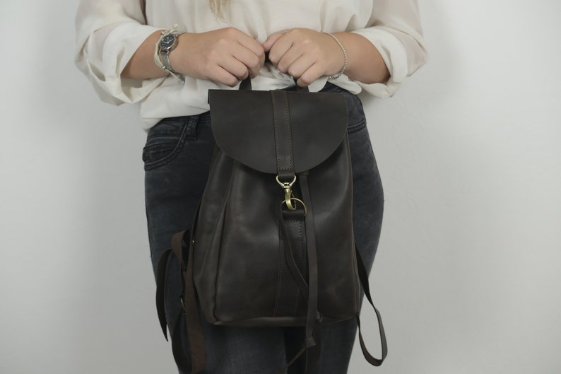 Pu&ograve; includere: Uno zaino in pelle marrone scuro con chiusura a patta ripiegabile fissata da un gancio in metallo color ottone e una chiusura ad anello. La borsa include spallacci regolabili e una maniglia superiore, con una finitura liscia e opaca esterna.