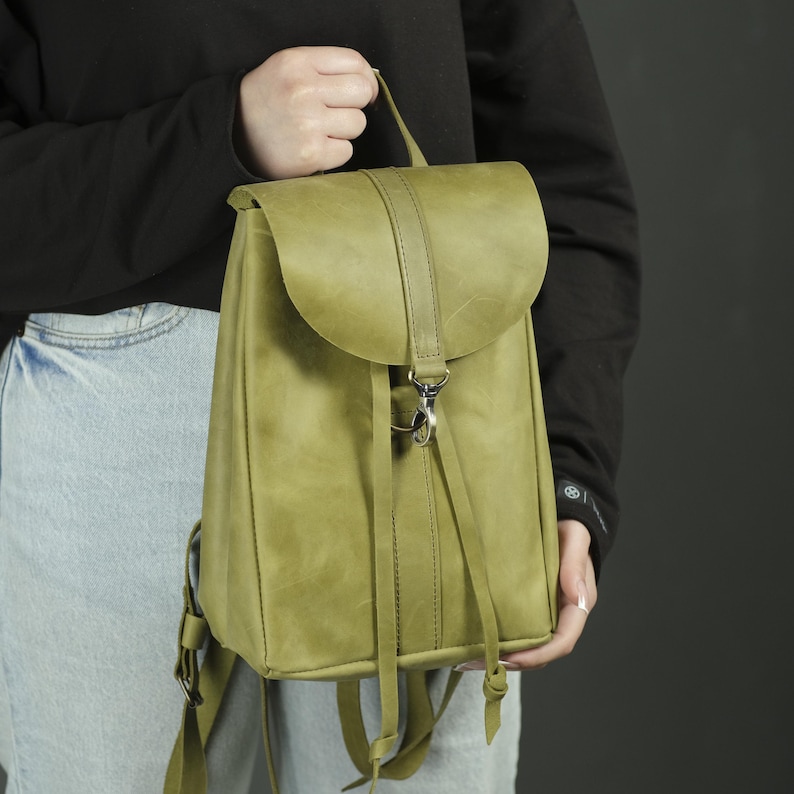 Pu&ograve; includere: Uno zaino in pelle verde salvia caratterizzato da una patta ripiegabile con cinturino verticale e chiusura con moschettone in metallo argentato. La borsa presenta lunghi lacci in pelle sul davanti e spallacci regolabili.