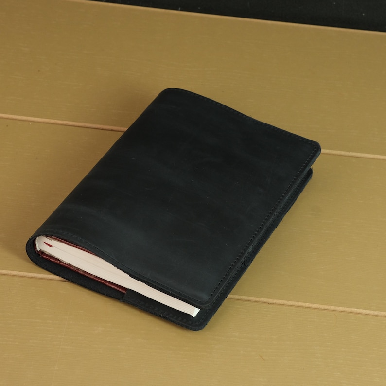 May include: A rectangular notebook encased in a matte black leather cover. The cover features visible stitching along the edges and a smooth, dark surface. The white pages of the notebook are partially visible along the bottom left corner edge.