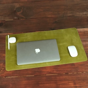 May include: A rectangular olive green leather desk mat featuring a closed silver laptop with an Apple logo, a white wireless computer mouse, a white stylus, and a small white charging case for wireless earbuds arranged on its surface.