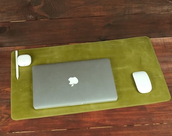 May include: A rectangular olive green leather desk mat featuring a closed silver laptop with an Apple logo, a white wireless computer mouse, a white stylus, and a small white charging case for wireless earbuds arranged on its surface.
