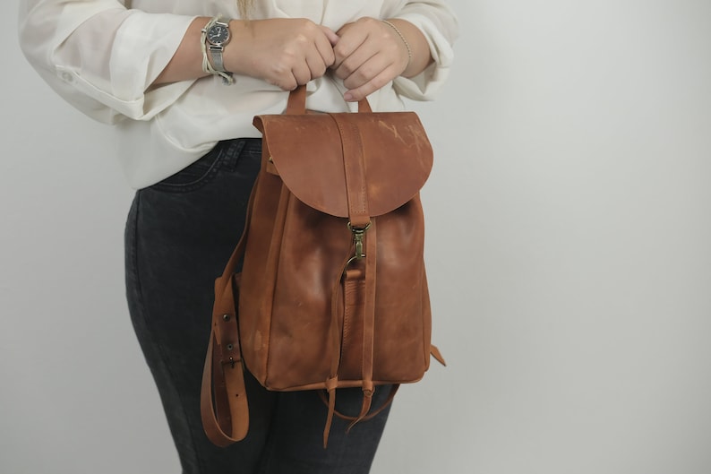 Pu&ograve; includere: Uno zaino in pelle marrone con patta ripiegabile dotata di chiusura a gancio in metallo color ottone, manico superiore e spallacci regolabili. La borsa presenta una finitura liscia e opaca con grana naturale visibile e lacci in pelle frontali.