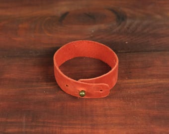 May include: A vibrant orange leather bracelet featuring a smooth, matte finish and a single metallic brass-toned stud closure. The band is constructed from a continuous strip of leather that overlaps at the ends to secure with the central metal stud.