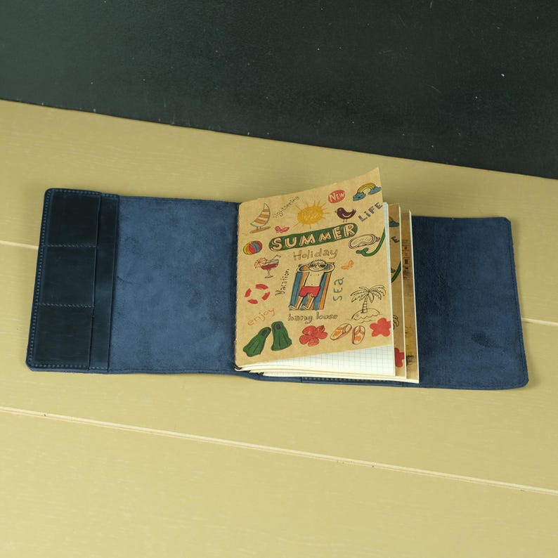 Pode incluir: Uma capa de caderno em camur&ccedil;a azul-escura com bolsos internos contendo um caderno de papel bege. A p&aacute;gina frontal do caderno apresenta ilustra&ccedil;&otilde;es de ver&atilde;o desenhadas &agrave; m&atilde;o e o texto "SUMMER LIFE Holiday".