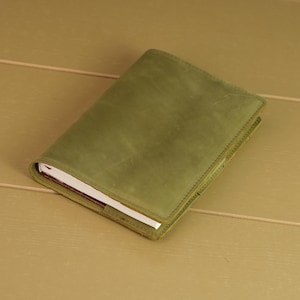 May include: A rectangular notebook encased in a matte olive green leather cover. The cover features visible stitching along the edges and a smooth, uniform texture. The white pages of the notebook are partially visible along the side edge.
