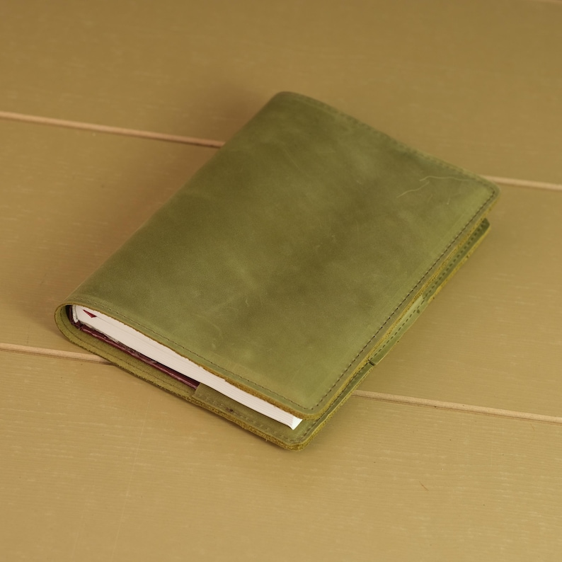 May include: A rectangular notebook encased in a matte olive green leather cover. The cover features visible stitching along the edges and a smooth, uniform texture. The white pages of the notebook are partially visible along the side edge.