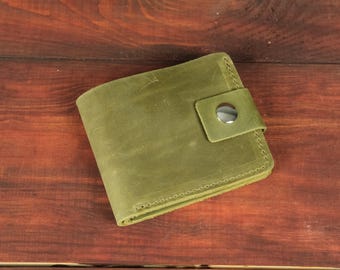 May include: A square olive green leather wallet featuring a smooth matte finish and a single strap closure secured with a polished silver-tone metal snap button. The wallet is constructed with visible tonal stitching along the edges.