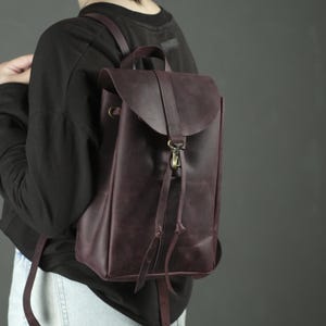 Pu&ograve; includere: Uno zaino in pelle viola scuro caratterizzato da una chiusura a patta con cinturino verticale centrale e un gancio in metallo color ottone. La borsa include spallacci sottili e due lunghi lacci in pelle che pendono dal meccanismo di chiusura.
