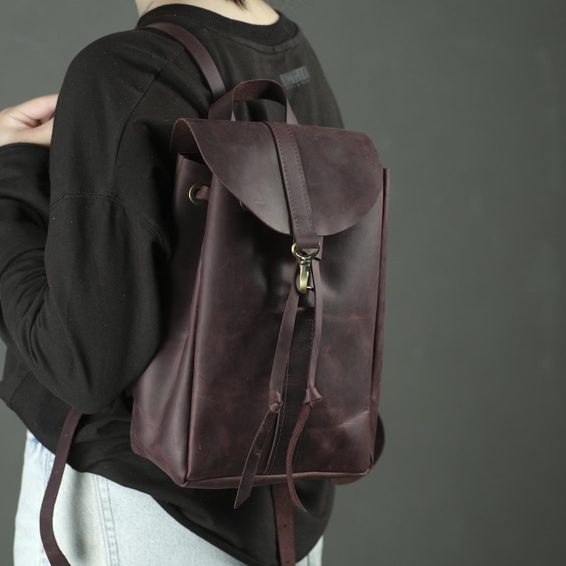 Pu&ograve; includere: Uno zaino in pelle viola scuro caratterizzato da una chiusura a patta con cinturino verticale centrale e un gancio in metallo color ottone. La borsa include spallacci sottili e due lunghi lacci in pelle che pendono dal meccanismo di chiusura.