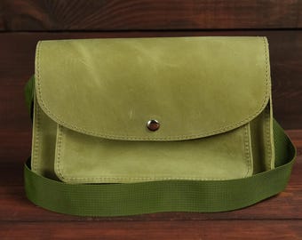 May include: A light olive green crossbody bag featuring a smooth matte finish, a rounded front flap with a single silver-toned metal snap button closure, a rectangular front pocket, and a wide matching olive green woven fabric shoulder strap.