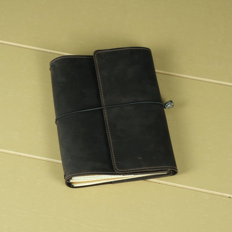 Pode incluir: Uma capa de di&aacute;rio em couro cinza-carv&atilde;o escuro com acabamento fosco e costuras vis&iacute;veis nas bordas. A capa &eacute; presa por um cord&atilde;o el&aacute;stico preto fino que envolve horizontalmente o centro, apresentando um pequeno detalhe de conta met&aacute;lica.